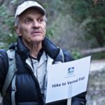 Close-up photo of Yosemite Conservancy naturalist and tour guide Dick Ewart holding a sign that reads "hike to Vernal Fall," with greenery visible behind him