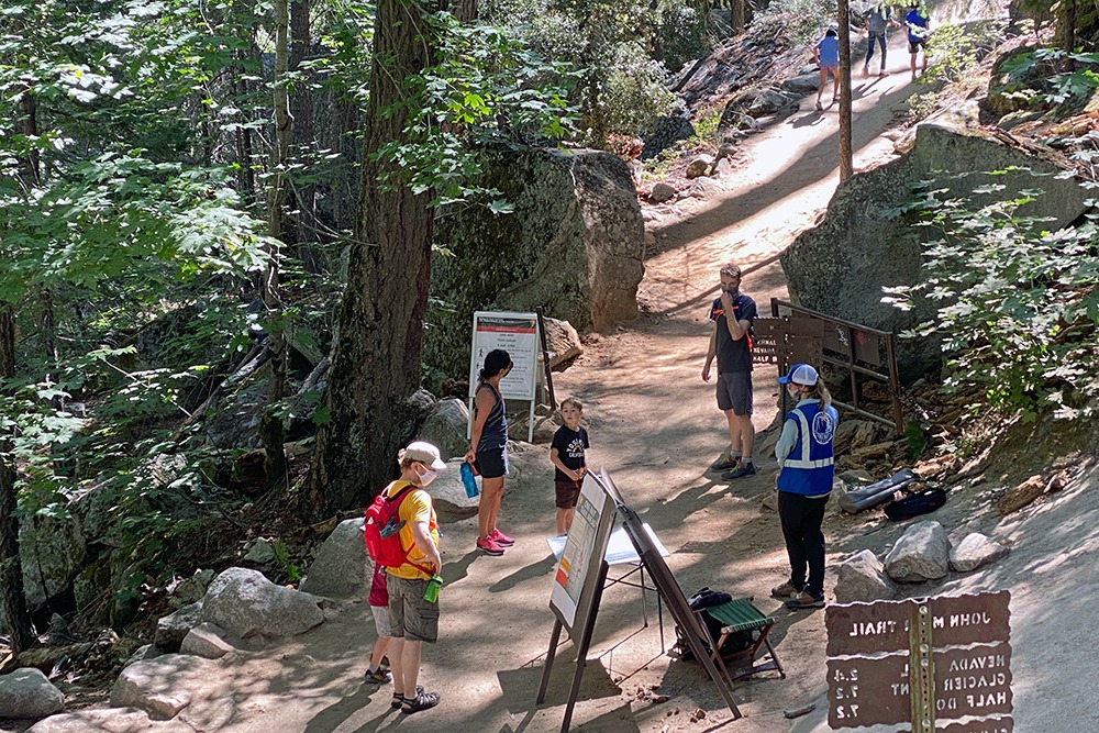 A group of hikers stands at the junction of the Mist and John Muir Trails in eastern Yosemite Valley, near several safety signs and a Conservancy naturalist in a blue Preventive Search and Rescue vest.