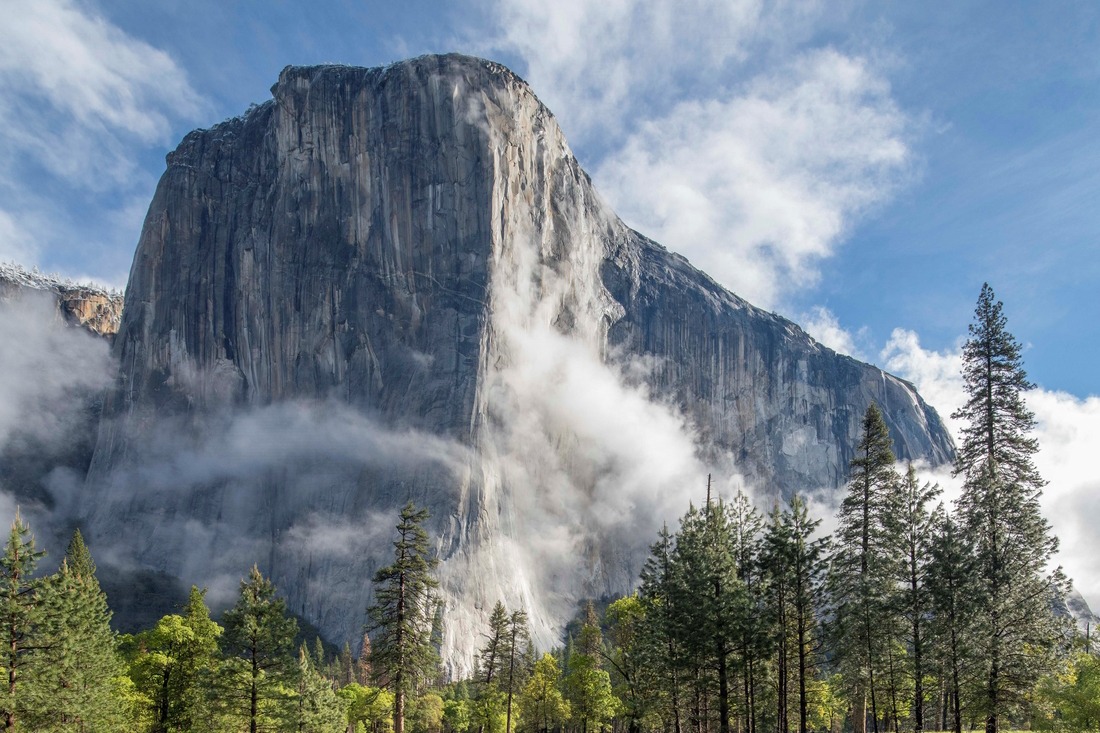 The granite walls of El Capitan behind wispy white cloud sand green trees. Photo by Matthew Scott.