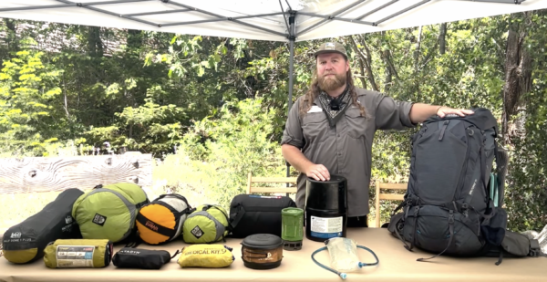 Marty stands at a table with backpacking gear laid out: packs, sleeping bags, first aid kits, etc are all spread out.