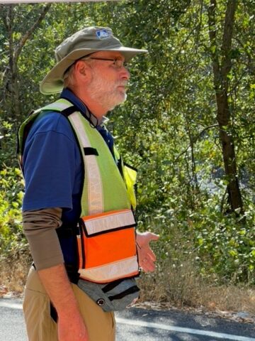Man standing with safety vest and broad rimmed hat directs the public along a trail in nature. 