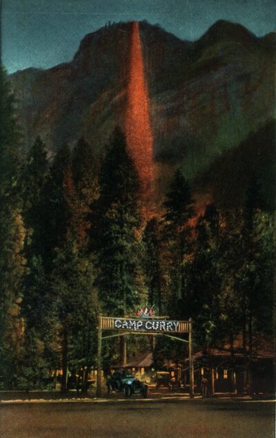 Looking at a dark night scene. Above there is a crest of a mountain and down the center there is a red orange glowing line of embers falling down the mountain. Below are trees and an archway that reads Camp Curry