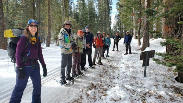 Group of bundled up adventurers with snow shoes standing in nature for a photo. Lots of smiles and conifers in the Yosemite winter scene. 