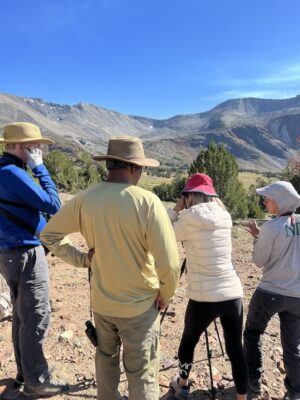 Group of people talking in the mountains. One person leans forward looking through a spotting scope hoping to see bighorn sheep.