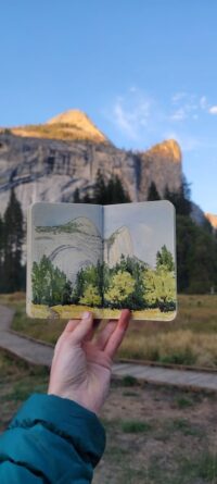 Artist's hand holding up sketchbook. The drawing of the mountain and meadow with trees matches the scene. 