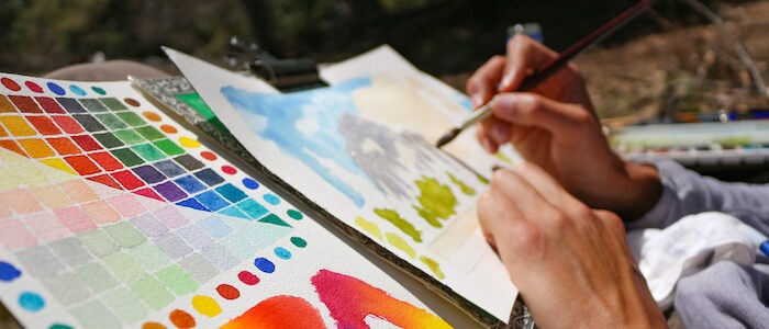 Artist workspace - two pieces of paper colored in painted colors, two hands at work with a brush in one hand.