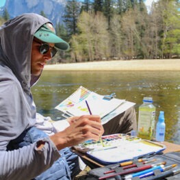 Artist in sun hoodie and hat painting by the river