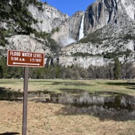 In the background a big white misty waterfall plummets over Yosemite walls in a series of cascades. In the foreground a sign reading Flood Water Level 11:00 p.m. 1/2/1997.