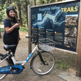 Woman standing with a Peet's coffee cup in her had. She is wearing a bike helmet and there is a blue bicycle before her. She is standing at a map of Yosemite Valley Trails decidind where to go on her summer day. 