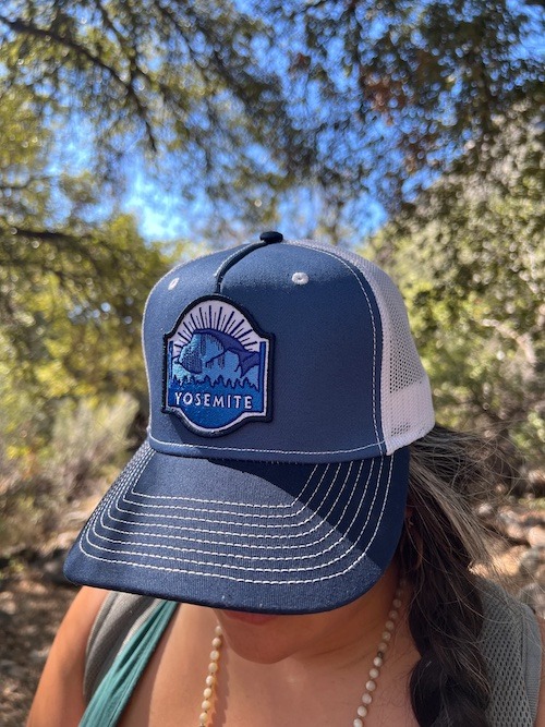 Looking at the blue Half Dome trucker hat. Navy Front with a Half Dome patch and then white mesh sides. The hat is sitting on a woman's head. She is standing in nature, surrounded by trees. 