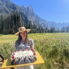 Adonia Ripple stands holding a large birthday cake in Sentinel Meadow to celebrate the Conservancy's 100th birthday to Yosemite in 2023. 