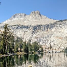 Looking across the glassy surface of May Lake, past a row of conifers on the left, up at the craggy peak of Mount Hoffman.