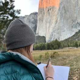 Woman creating art outside, curled up in a beanie and puffy drawing El Capitan at the last light of day. 