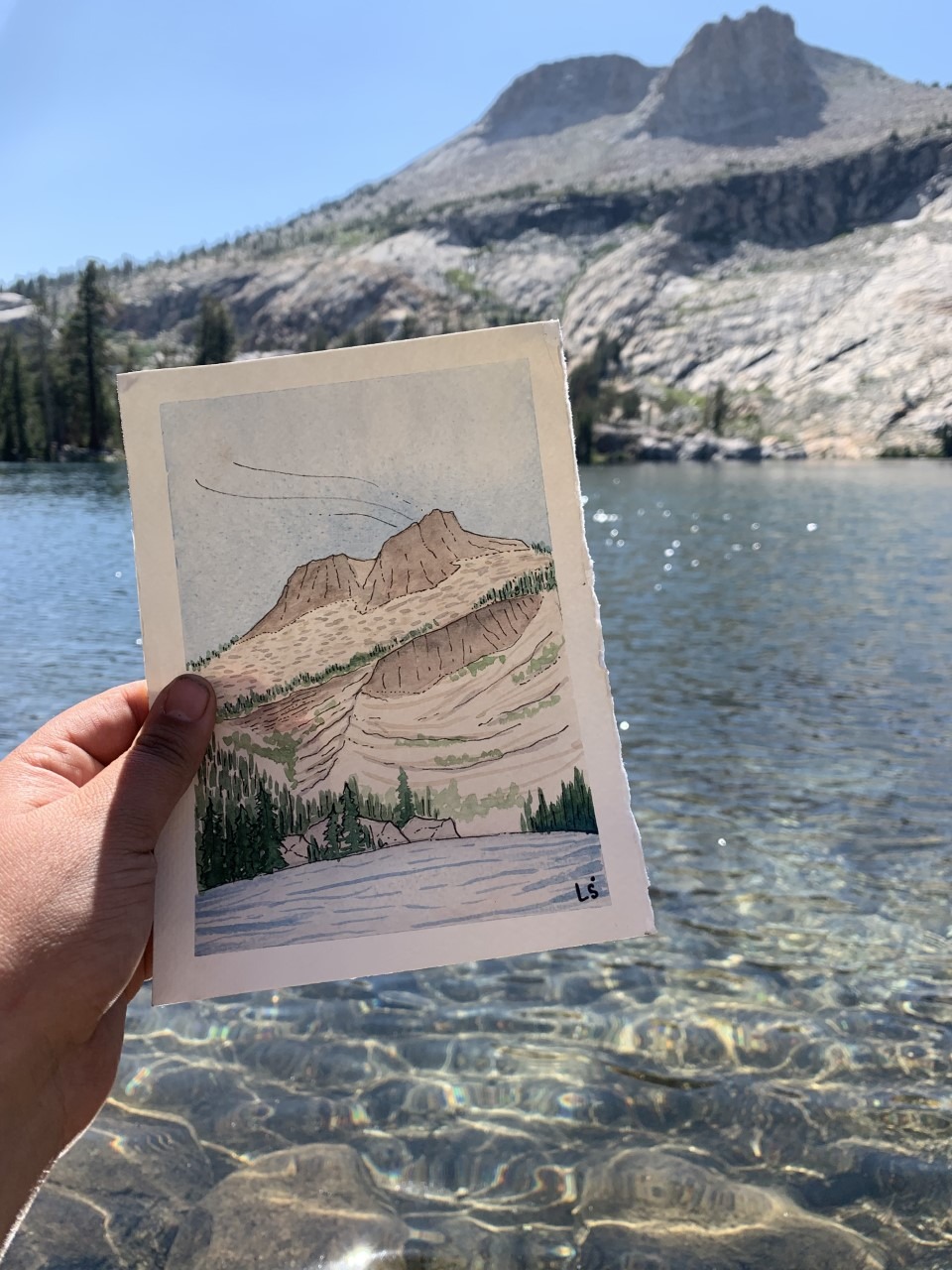 photo of a person holding up a painting of mount hoffman in front of mount hoffman and glimmering water