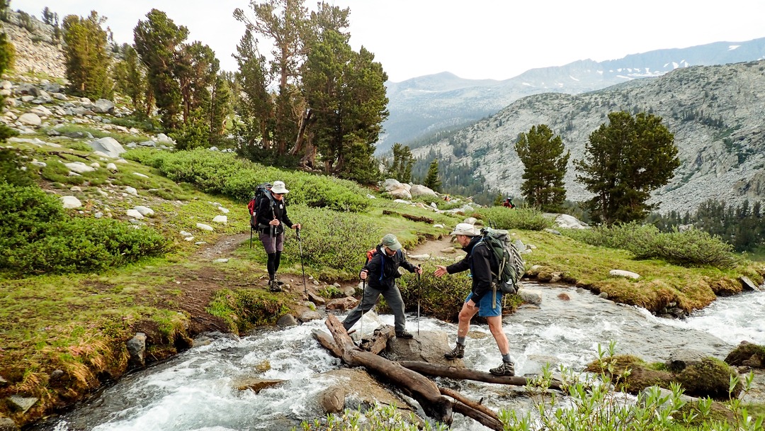 Three backpackers cross a stream in an open grassy area. Two backpackers stand on either side of the stream, while the third attempts to cross using rocks and logs in the middle of the stream. In the background are beautiful mountain ranges.