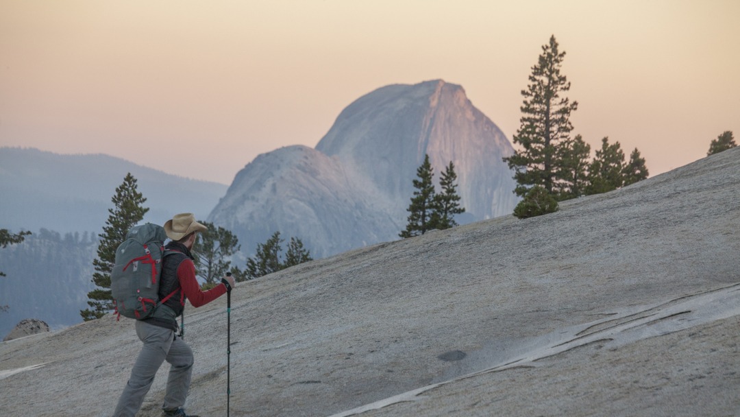 Man pictured backpacking uphill with half dome in the background. 