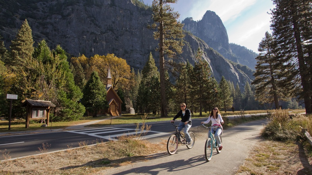 Two adults bike on a paved path in Yosemite with the Yosemite Valley Chapel, trees, and large rock formations in the background.