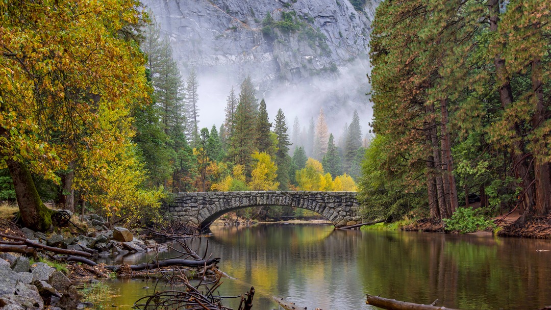 A stone bridge crosses a clear river. The surrounding trees have their fall colors and there is fog in the background.