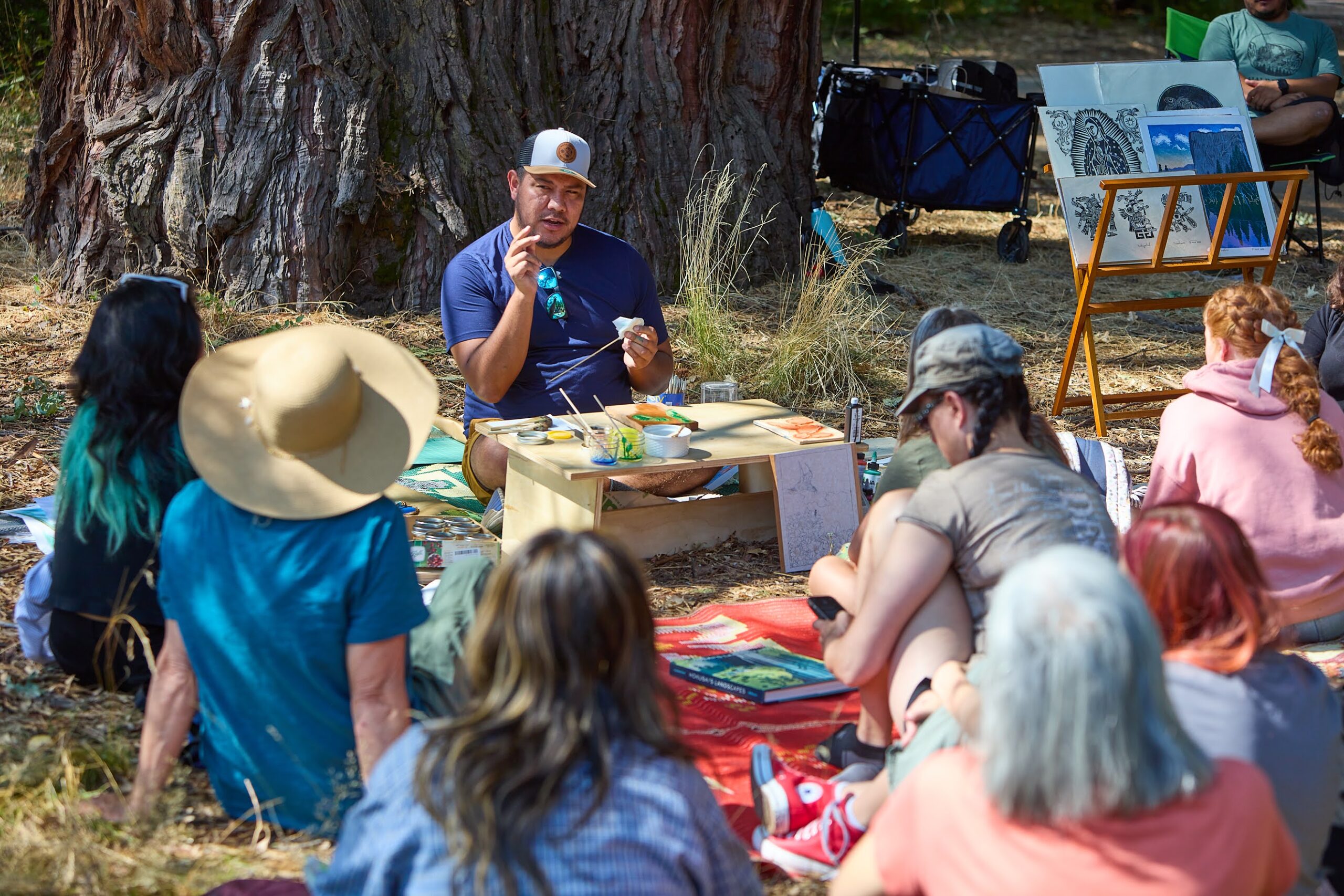 photo of someone sitting on the ground with many others. the person in the front is teaching an art class.