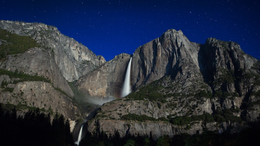 Yosemite Falls and the valley below aglow from starlight and moonlight. Above the falls, the night sky is dotted with stars. Yosemite Valley is a popular place for stargazing in Yosemite.