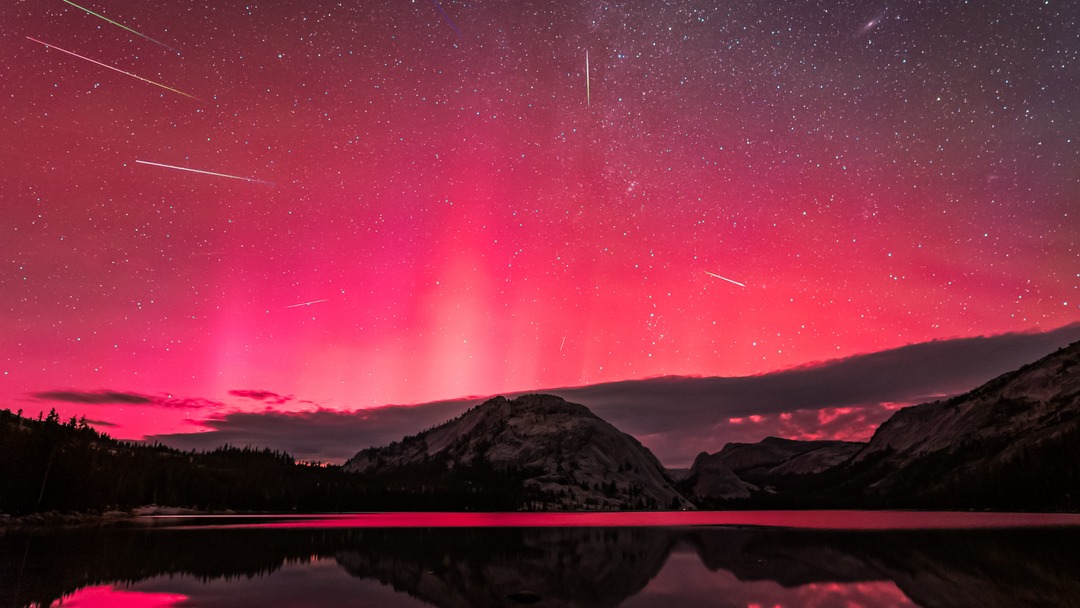 A deep pink sky filled with shooting stars reflects in a lake below. In the distance, peaks and forest can be sen on the lake's shore.