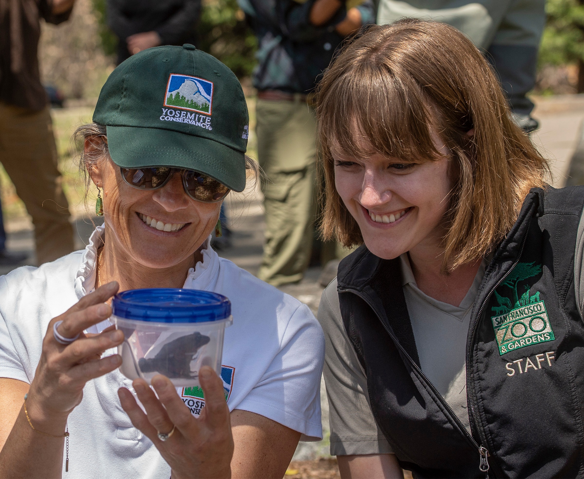 Two people smile at a frog in a plastic container.