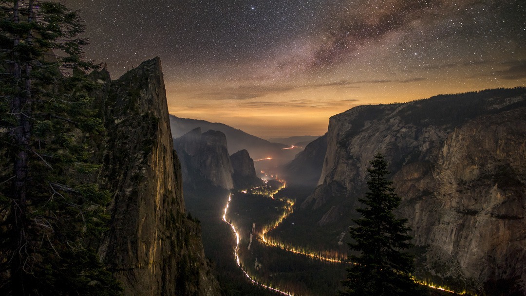 Car lights dot the floor of a deep valley at night. On either side of the valley are trees and tall cliffs.