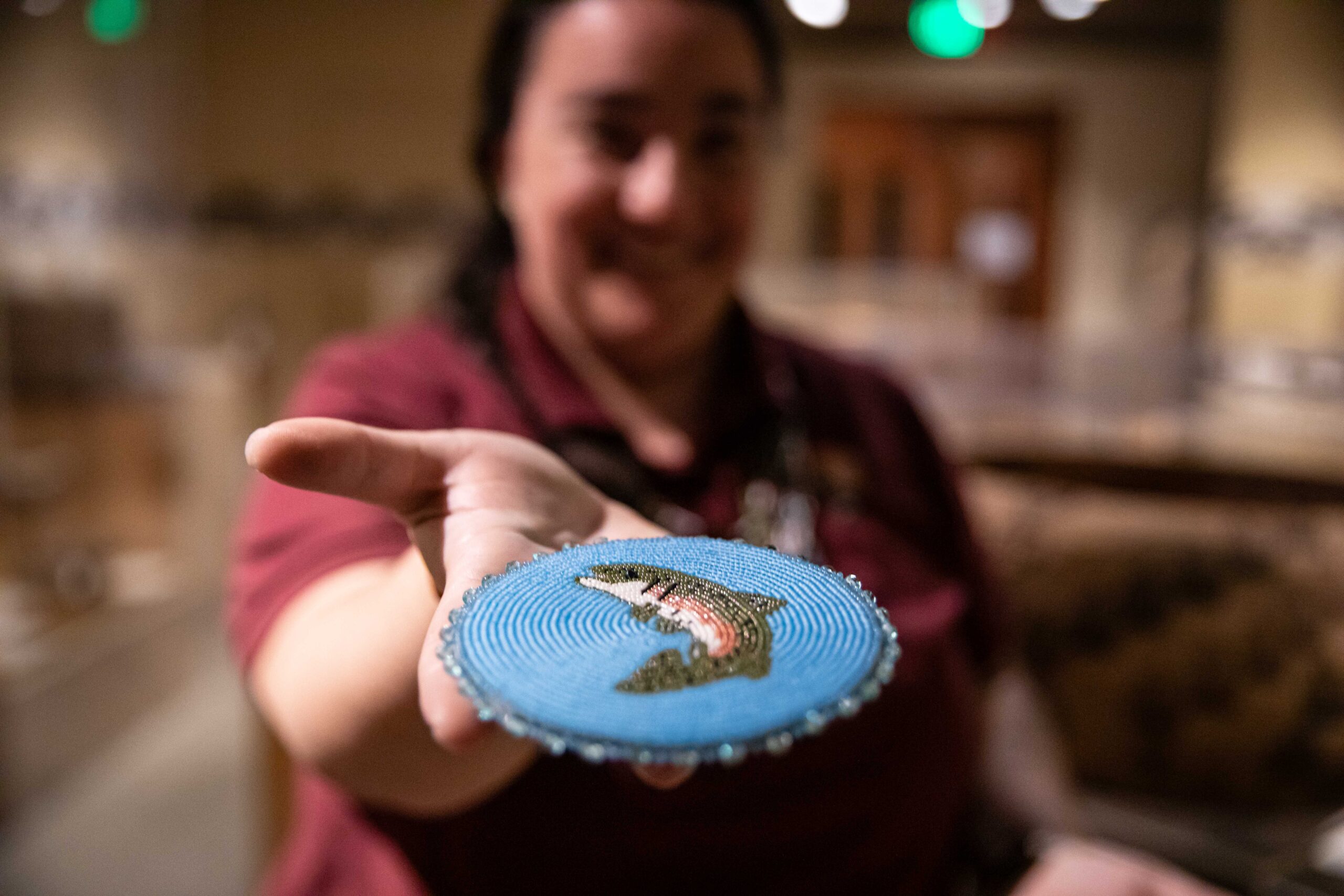 Woman holds beaded medallion necklace that is blue with a green fish on it. We are still here.