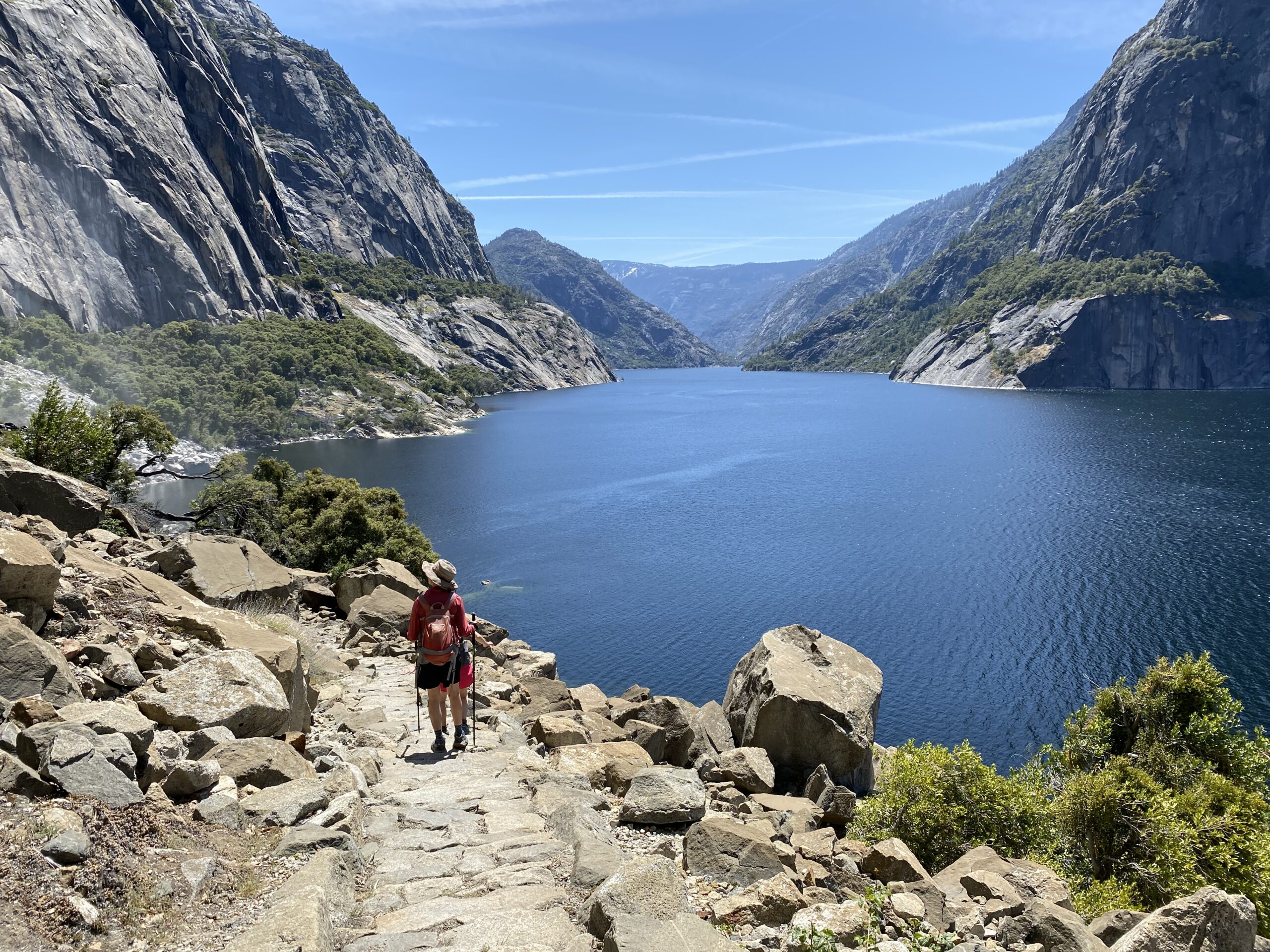 A single hiker walks away from the camera alongside the Hetch Hetchy reservoir.