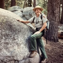 Youthful Dick Ewart during his ranger years poses next to a boulder in ranger uniform.