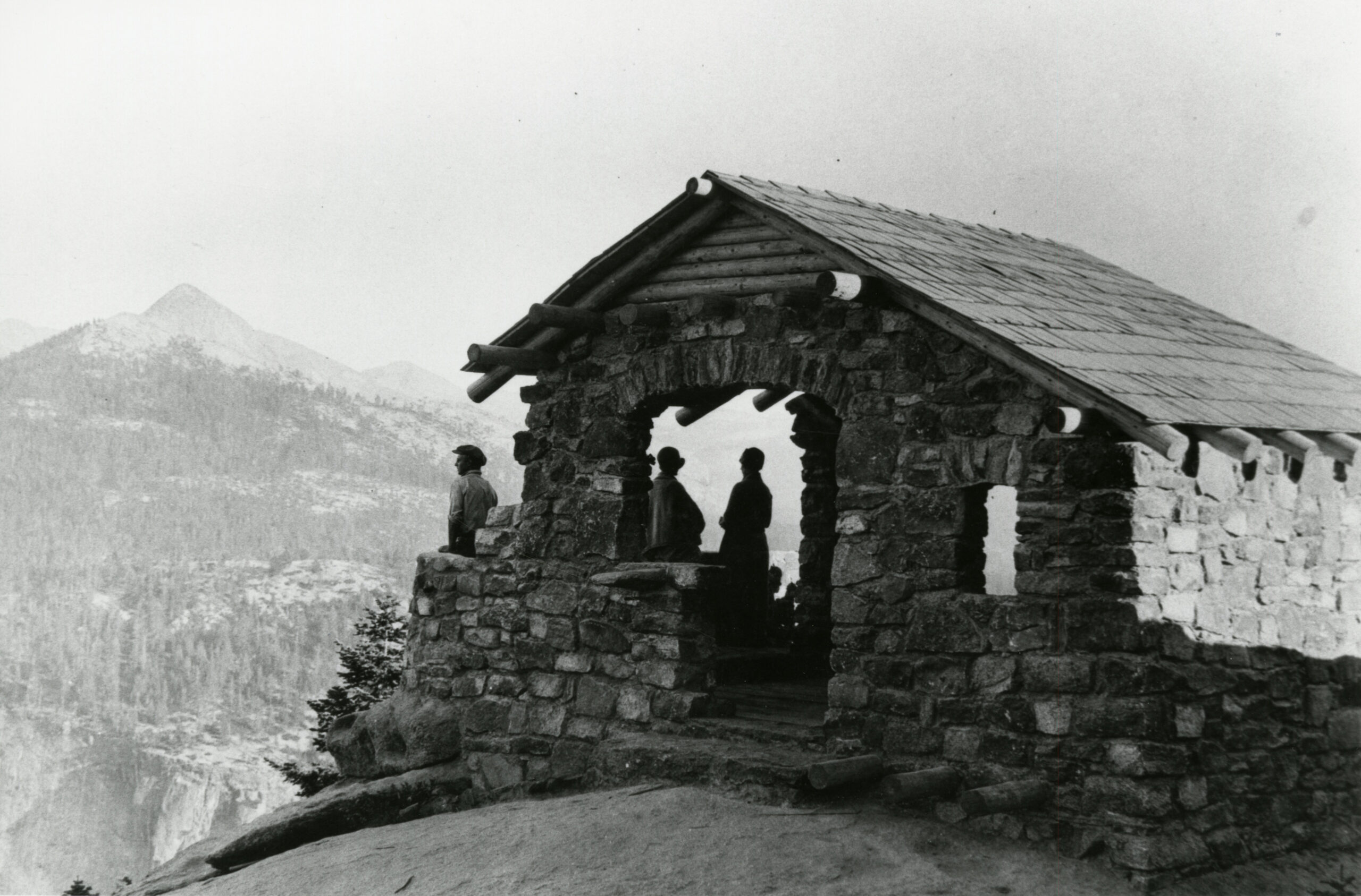 Historical, black and white photo of the stone Geology Hut at Glacier Point. Three individuals stand inside the hut, gazing out over the valley bellow.