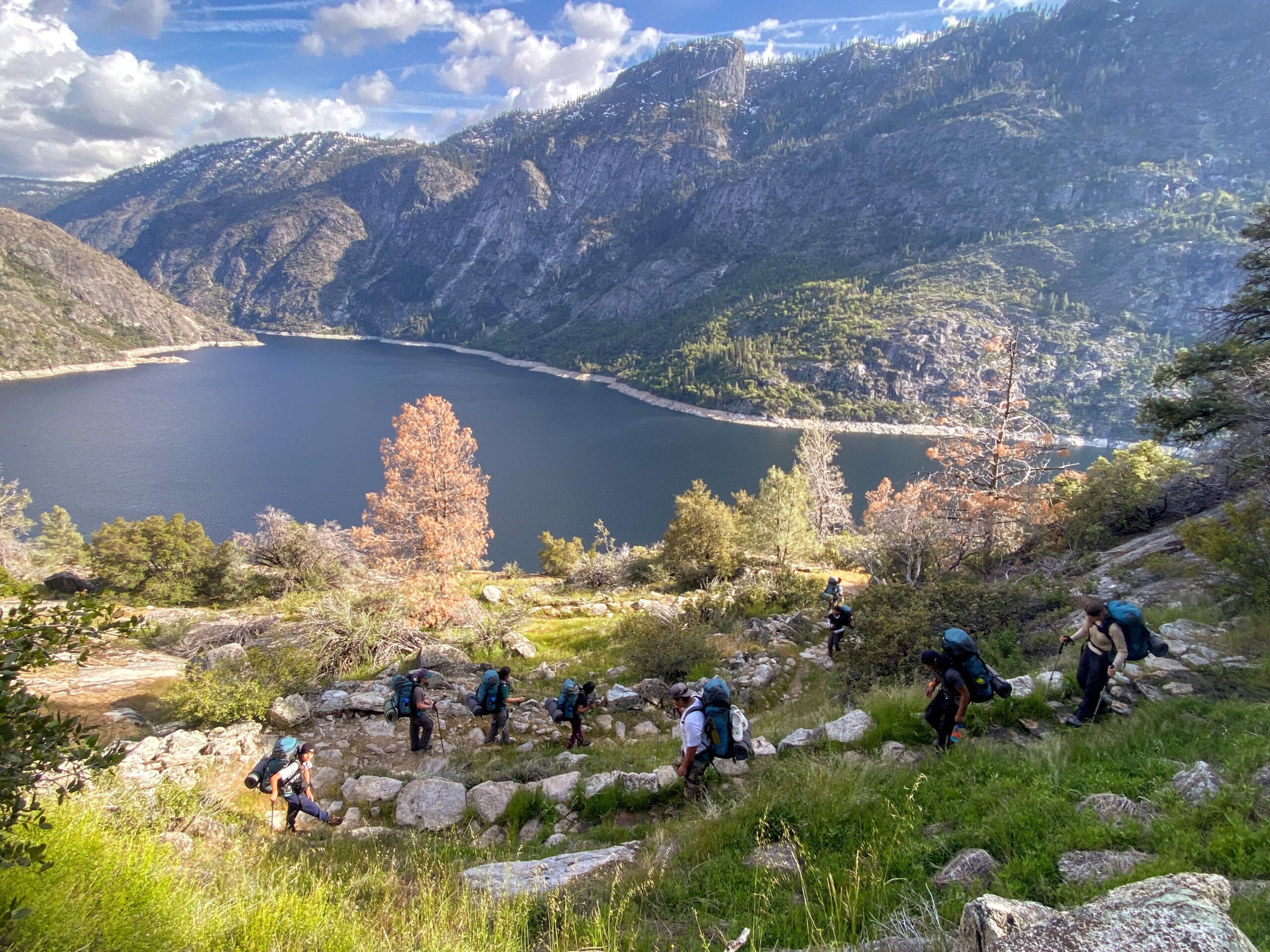 Young people hike down a grassy hillside towards a large lake that's surrounded by granite cliffs in Yosemite in 2025.