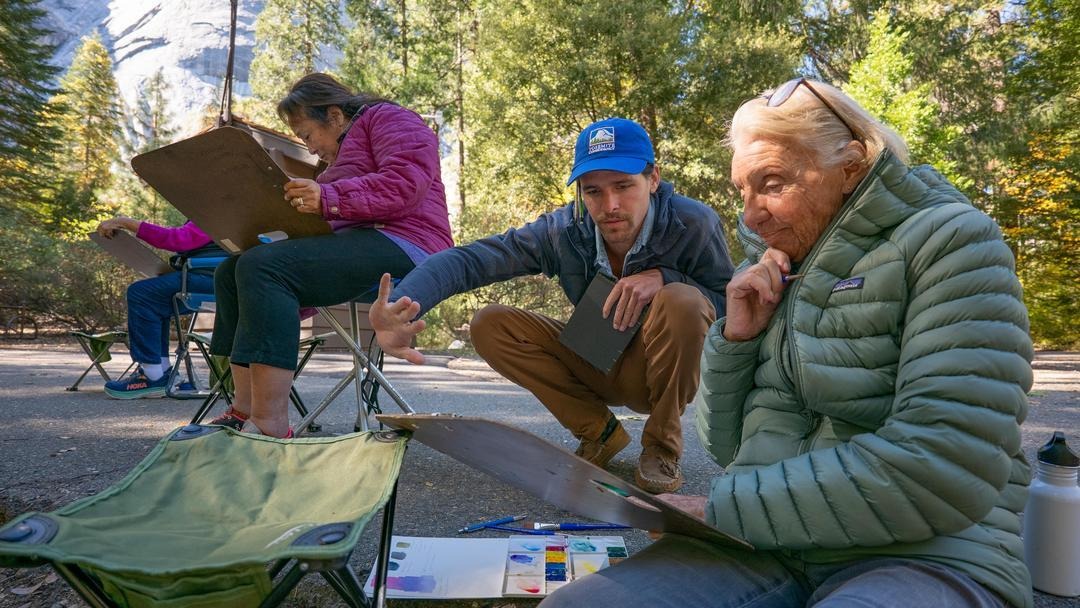 Two people sit on the ground. One has blonde hair, is wearing a green puffer jacket, and is working on an art piece. The other wears a blue hat, blue jacket, and tan pants, and is helping the artist with her piece. In the background, two other people wear pink/purple jackets and work on art their own art piece among trees.