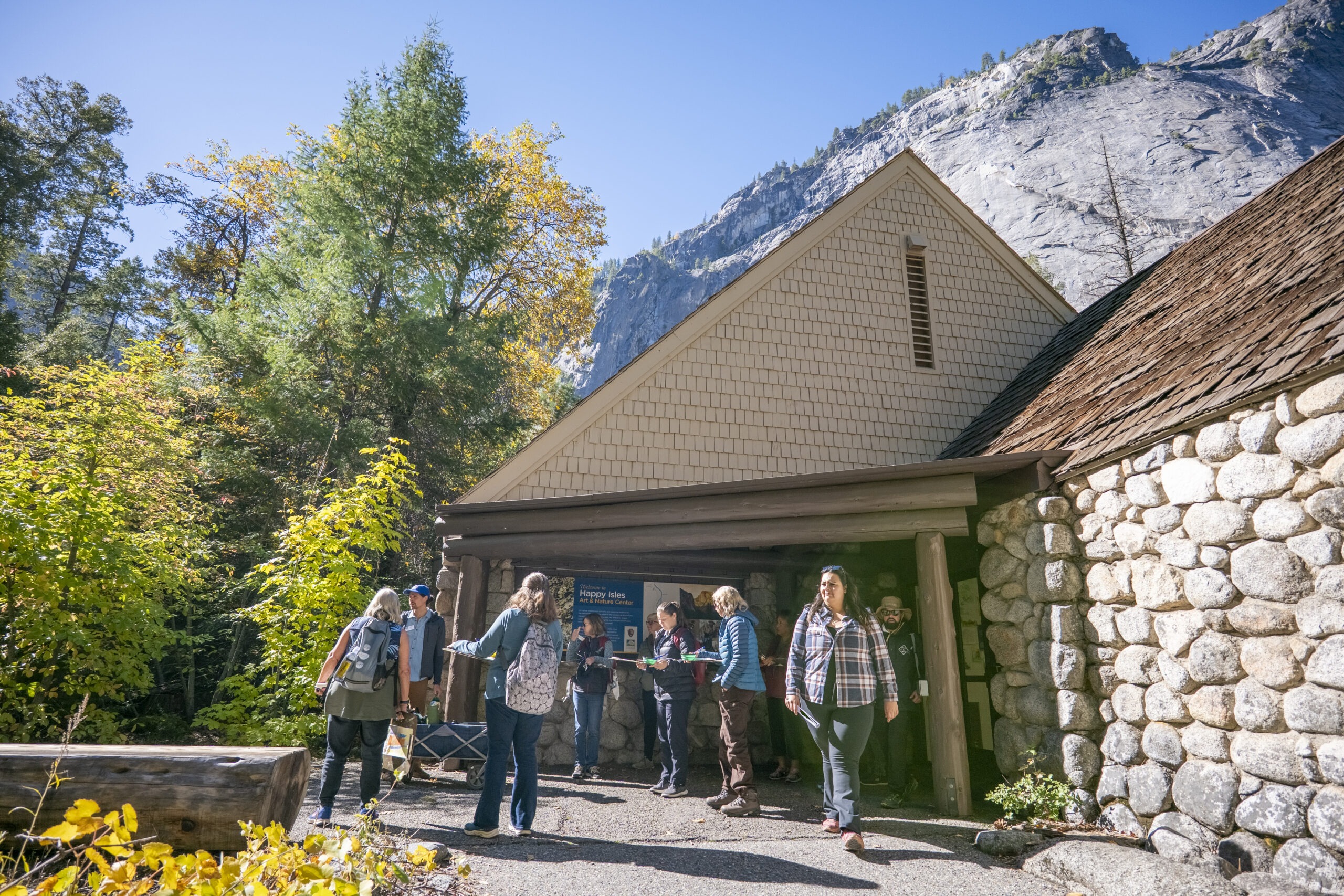 People stand outside a stone building surrounded by fall foliage and towering granite rocks.